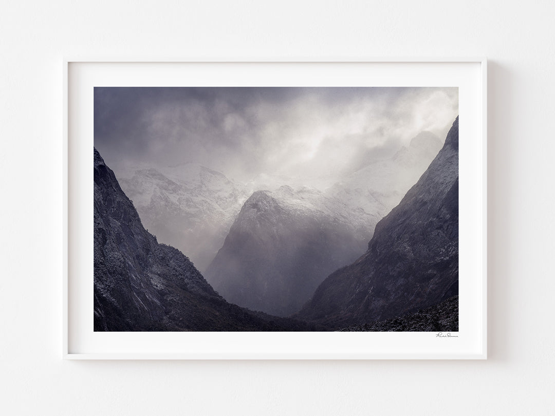 A framed landscape art print depicting a misty mountain valley with snow-capped peaks, mounted on a clean white wall.