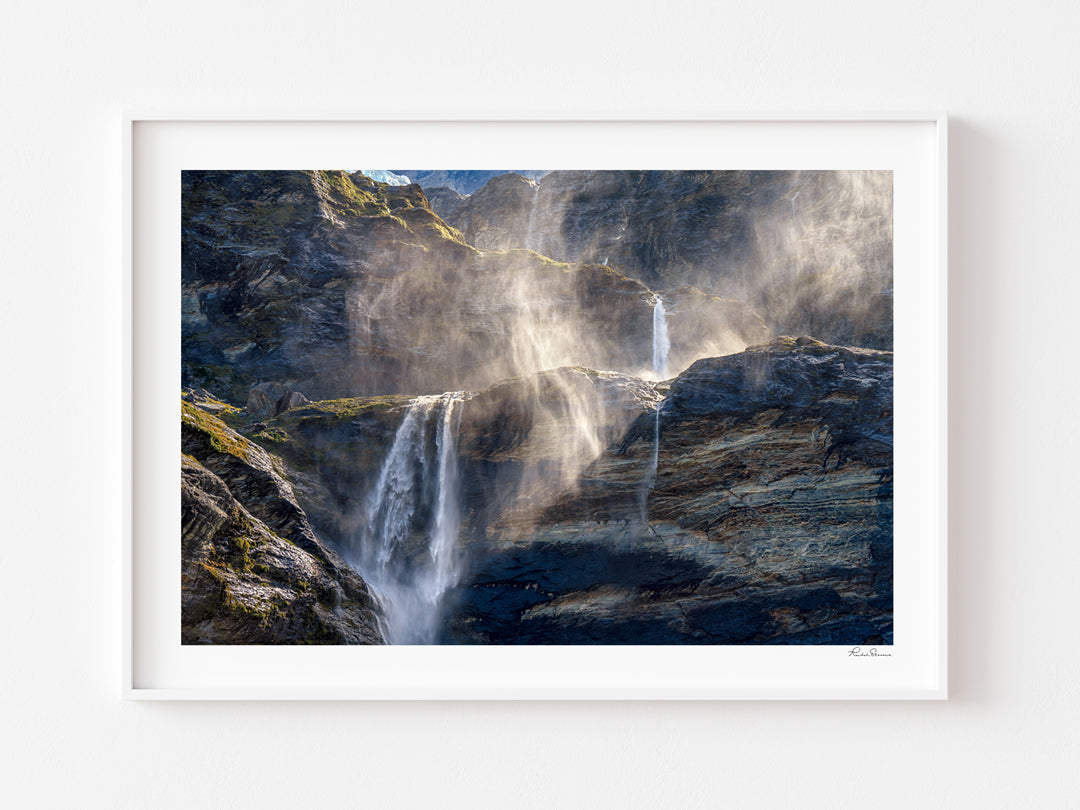 A framed landscape photograph of a waterfall cascading down dark rocky cliffs with mist, presented in a white frame against a clean white wall.