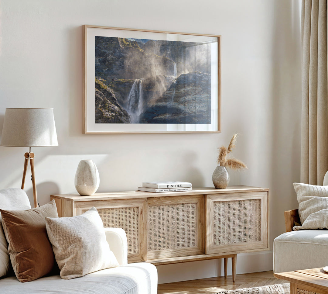 A framed landscape art print depicting a waterfall in a forest, styled in a modern living room above a wooden sideboard.