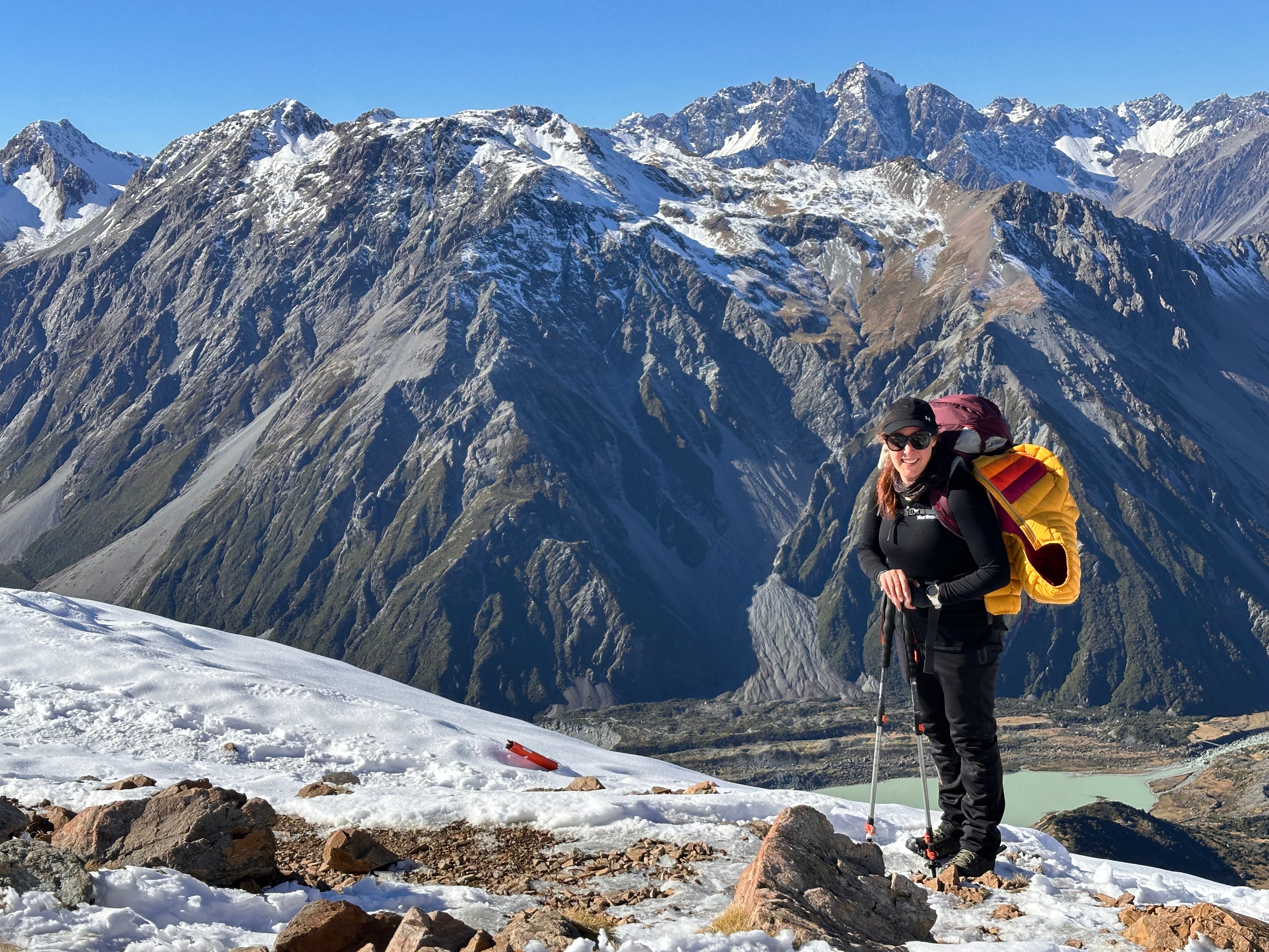 A hiker stands on a snowy mountain ridge with a dramatic backdrop of rugged snow-dusted peaks and a blue sky. The person is wearing black outdoor gear and carrying a large yellow and red backpack.