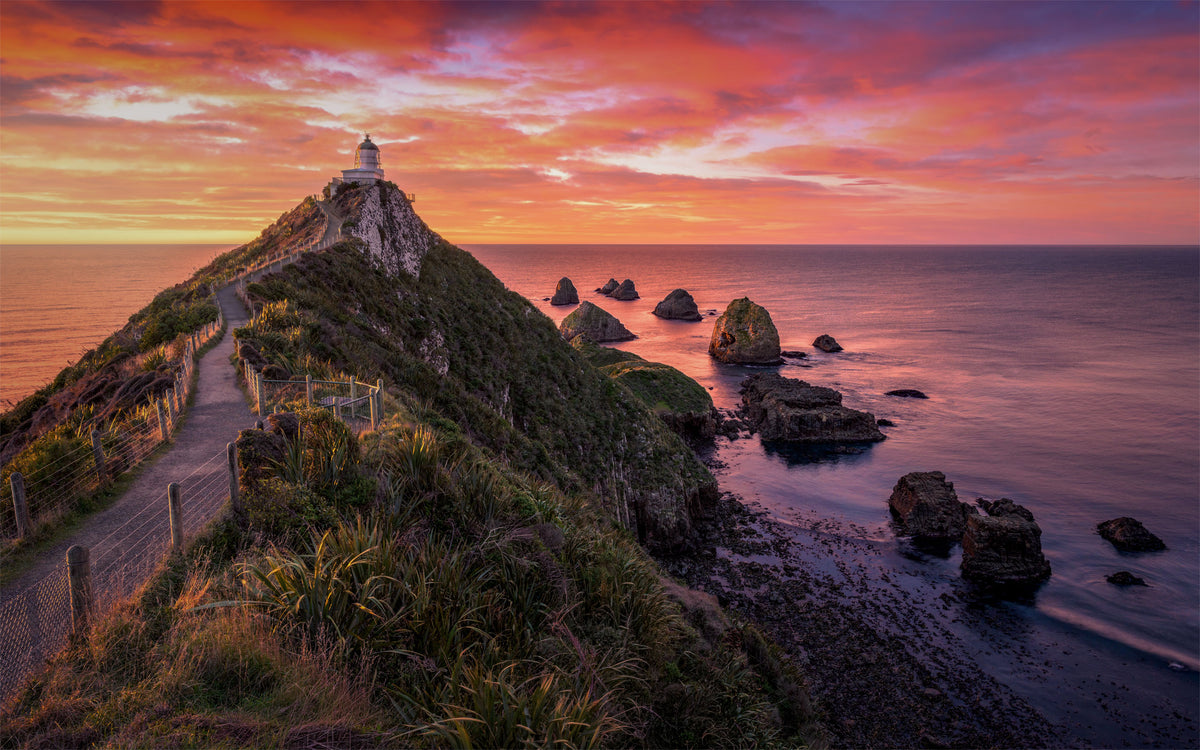 A dramatic coastal landscape at sunset with a lighthouse atop a grassy cliff, a winding path along the edge, rocky sea stacks, and a vivid orange, pink, and purple sky over the ocean.