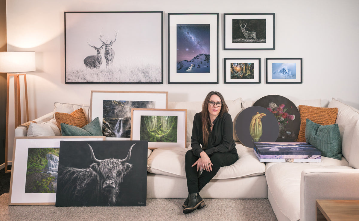 A woman sits on a white sofa in a styled living room, surrounded by multiple framed and matted art prints and paintings, including a large black-and-white Highland cow, a deer, a night sky, and floral still life.