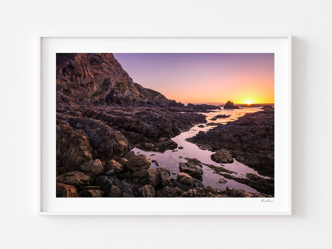 Rocky coastline at Whiterea Park at sunset, displayed in a white frame with a white mat.